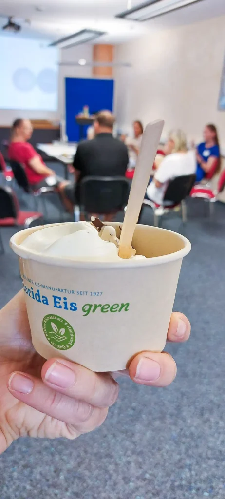 Im Vordergrund hält eine Hand einen kleinen Becher mit hellem Eis und einem Holzlöffel. Auf dem Becher steht „Florida Eis green“ sowie ein grünes Siegel mit den Begriffen Klimaschutz, Handwerk und Qualität. Im Hintergrund sitzen mehrere Personen.